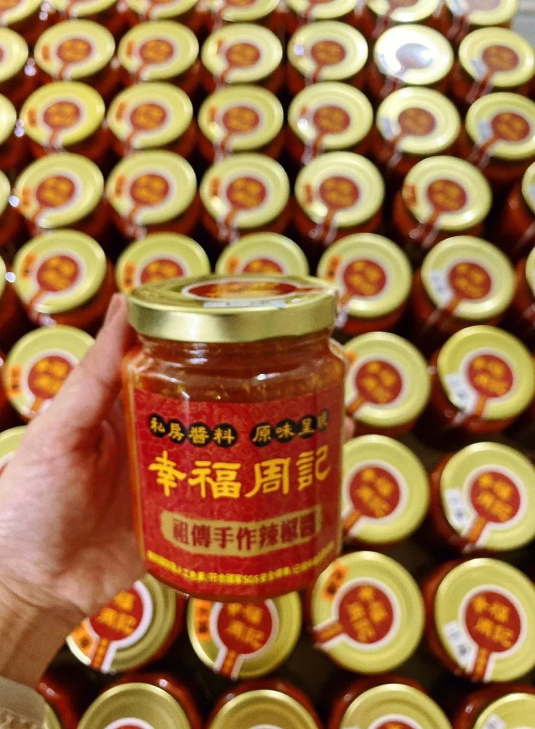 手拿幸福周記辣椒醬,後方整齊排列多瓶產品